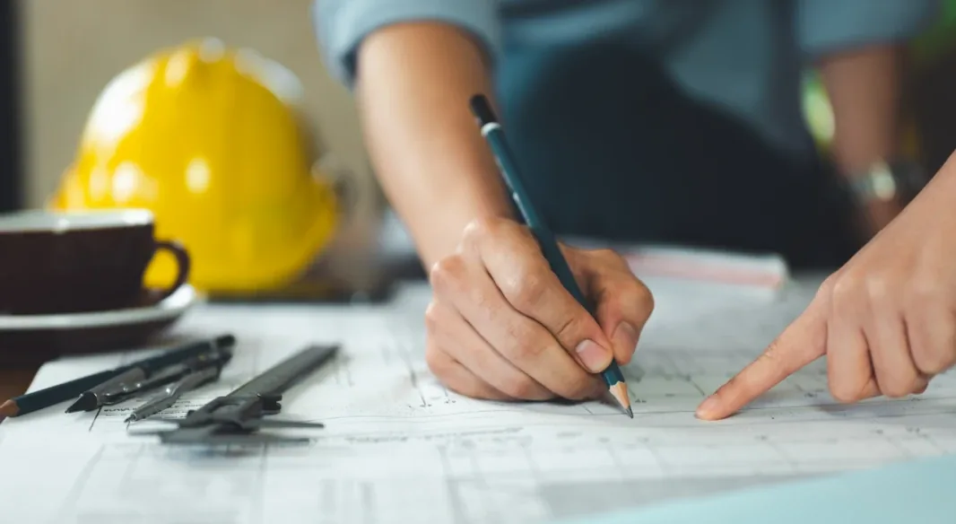 Engineer marking up structural drawings at a desk beside a hard hat